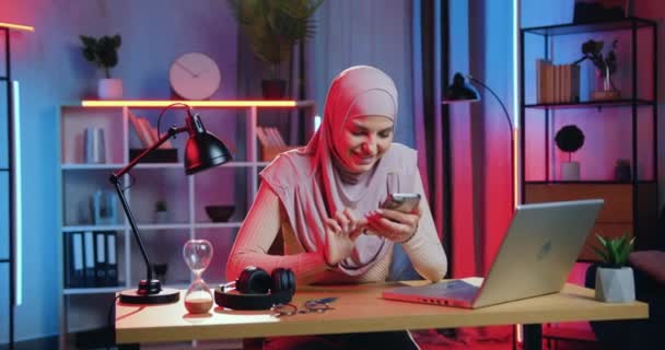 Portrait de sympathique souriant jeune femme musulmane heureuse en foulard qui assis à son lieu de travail et bavarder avec des amis lors de la pause dans son travail sur ordinateur portable à la maison le soir — Video