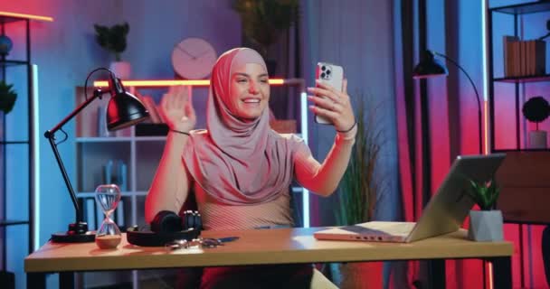 Beautiful happy smiling modern young muslim woman in traditional hijab enjoying pleasant video chat with friend or family on beautifully lighted room in the evening — Stock Video