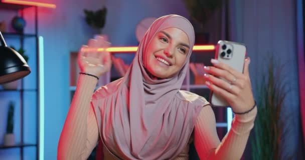 友達や家族とビデオチャットを介して話しているヒジャーブの美しい笑顔30歳のアラビア語の女性が夜に自宅でモバイルで — ストック動画