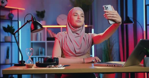 Bedårande glad glad leende ung arabiskt kvinna i traditionell slöja sitter på sin arbetsplats på kontoret på kvällen och gör selfie på smartphone — Stockvideo