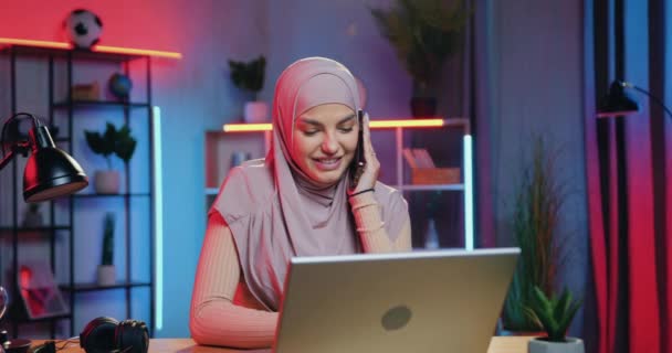 Aantrekkelijk verrast vrolijke jonge Arabische vrouw krijgt goed nieuws op laptop wanneer zitten op haar werkplek thuis in de avond en praten op mobiele — Stockvideo