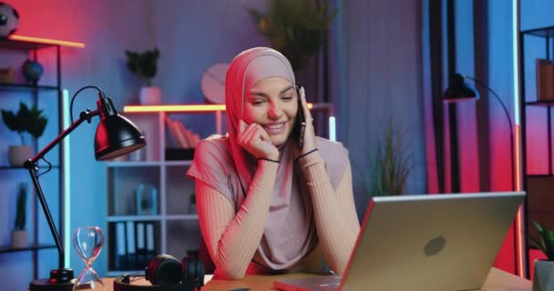 楽しい電話会話をしながら、夜に自宅でコンピュータ上で彼女の仕事を中断するスカーフで楽しい笑顔のんきな若いムスリム女性の肖像画 — ストック動画