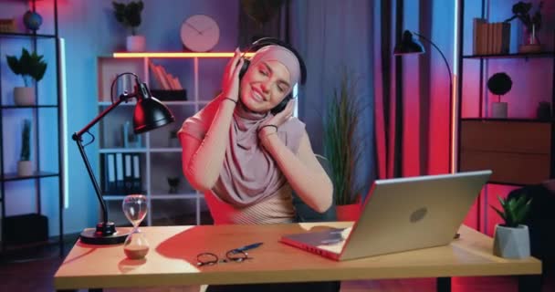 Lovely puas tersenyum bahagia muda arabian wanita dalam jilbab memakai earphone dan menggerakkan tubuhnya dalam irama musik favorit di malam hari di rumah — Stok Video