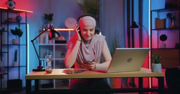 Fritidskoncept där attraktiv leende avslappnad ung muslimsk kvinna i slöja njuter av favoritmusik i hörlurar från telefon spellista hemma på kvällen — Stockvideo