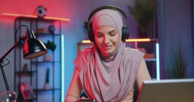 Gülümseyen mutlu, rahat, hafif menekşeli başörtüsü takmış, güzel dekore edilmiş, renkli ışıklandırmalı odada güzel müzikler dinleyen 30 'lu yaşlarda Arap bir kadın.