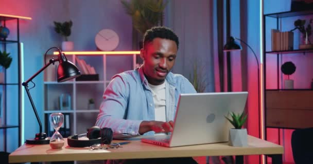 Красавчик улыбается, уверенный в себе опытный молодой африканский американец доволен, когда закончил свой компьютерный проект дома и опираясь на стул положить руки за голову смотреть на экране — стоковое видео
