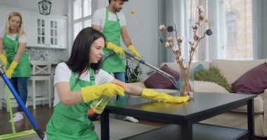 Yeşil önlüklü ve koruyucu eldivenli, gülümseyen, çalışkan bir kadının ön görüntüsü. Mikrofiber bez ve deterjanla masayı silerken temizlik ekibinin diğer üyeleri odayı temizliyor.