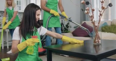 Temizlik şirketinden güzel, neşeli genç bir kadın mikrofiber bez ve deterjanla masayı siliyor, başparmaklarıyla kameraya bakıyor ve diğer meslektaşları da müşterilerin evini süpürüyor.