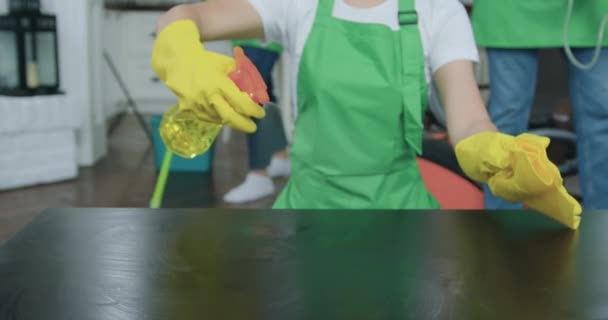 Неизвестная женщина распыляет чистящие средства на поверхность столов и вытирает их микроволоконной тканью во время уборки дома клиента вместе с другими членами службы уборки — стоковое видео