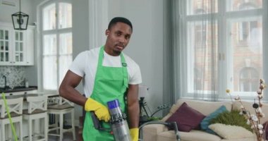 Yakışıklı, kendine güvenen Afro-Amerikan üniformalı genç bir temizlikçi. Müşteri evini elektrikli süpürgeyle temizlerken kamerada poz veriyor.