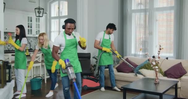面白い若いアフリカ系アメリカ人の清掃サービスの労働者がカーペットを掃除し、ヘッドフォンで音楽の下で踊り、ギターのように真空で演奏する一方、清掃チームの他のメンバー — ストック動画