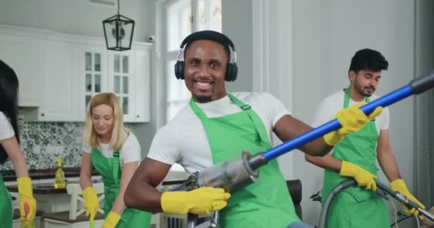 他のメンバーと一緒に顧客の家を掃除しながら、ギターのように掃除機で遊んでヘッドフォンでハンサムな陽気な若い黒い肌のクリーナー — ストック動画