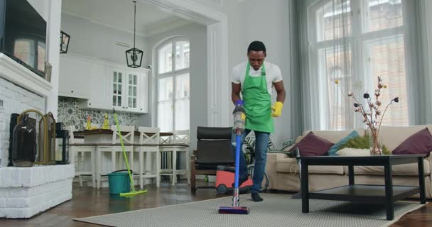 Симпатичный, веселый, профессиональный чернокожий работник уборки, занимающийся мокрой уборкой с пылесосом и танцующий во время работы в квартире клиента — стоковое видео
