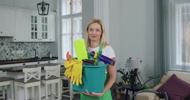 Porträtt av snygg leende vuxen blond kvinna från rengöringsservice som håller i hennes händer hink med olika rengöringsmedel och tittar på kameran under rengöring comntemporary flat — Stockvideo