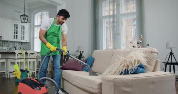 Schöne positive qualifizierte unrasierte Kerl in Uniform der Reinigung Service Staubsaugen bequemes Sofa während der Aufräumarbeiten zeitgenössischen Haus — Stockvideo