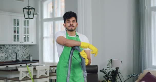 Beau professionnel hautement qualifié jeune homme barbu-travailleur du service de nettoyage appuyé sur l'aspirateur et regardant la caméra tout en nettoyant la maison moderne — Video