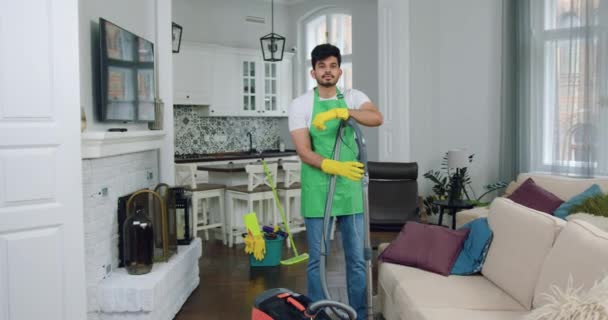 Müşterilerin evindeki işi sırasında kameraya elektrikli süpürgeyle poz veren iyi huylu, kendine güvenen, yetenekli bir temizlik işçisi portresi. — Stok video