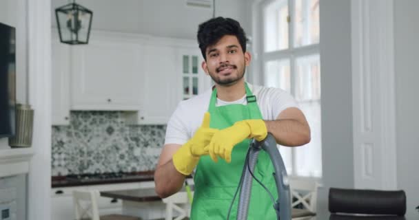 Atraente sorridente jovem limpador profissional em pé uniforme com aspirador de pó na frente da câmera e mostrando gesto ok ao limpar apartamento bem decorado — Vídeo de Stock