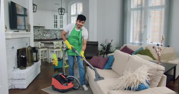 Pleasant smiling confident 30-aged bearded guy-cleaner in uniform vacuuming soft couch in modern flat and looking at camera with lucky face,cleaning service concept — Stock Video