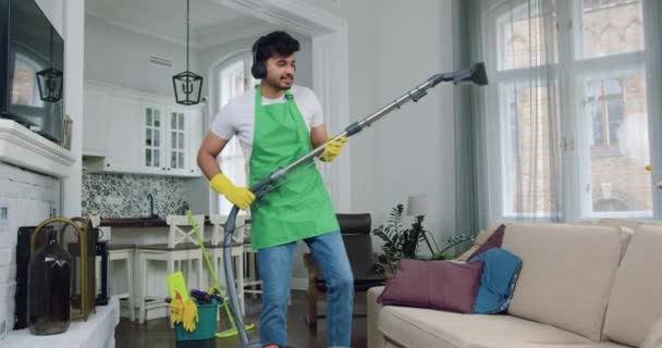 Portrait of likable joyful professional young cleaner in uniform which listening music in headphones while vacuuming sofa dust mites during cleaning up clients flat — Stock Video