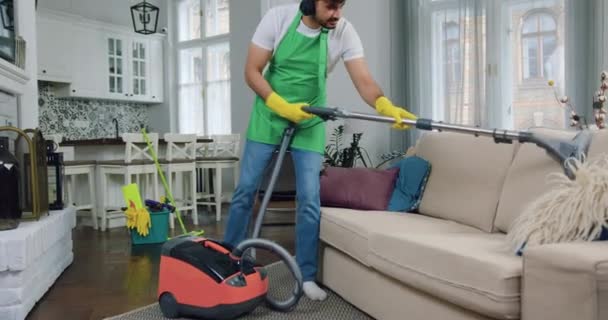 Likable positive smiling skilled bearded worker of cleaning service in headphones vacuum uming comfortable sofa during cleaning up contemporary flat — стоковое видео