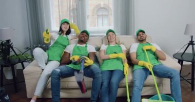 Müşteri odasının oturma odasındaki temizlik malzemeleriyle kameraya poz veren mutlu, çok ırklı temizlik personeli.