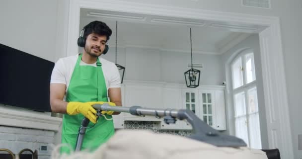 Agradable sonrisa contenta profesional barbudo limpiador en uniforme disfrutando de la música moderna en los auriculares mientras aspira sofá doméstico en piso contemporáneo — Vídeos de Stock