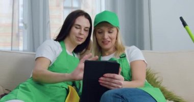 Oturma odasındaki yumuşak kanepede dinlenen güzel, mutlu ve deneyimli kadın temizlikçiler. Müşteri evini temizleme zamanı geldiğinde tablet bilgisayar kullanıyorlar.
