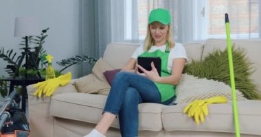 Çekici, yeşil önlüklü ve şapkalı kendine güvenen sarışın kadın rahat koltukta oturuyor ve evini temizledikten sonra tablet bilgisayar kullanıyor.