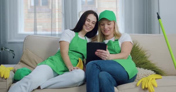 Улыбающиеся довольные молодые женщины-уборщицы отдыхают на диване в современном доме и смотрят забавные видео на планшетном компьютере во время перерыва во время уборки квартиры. — стоковое видео