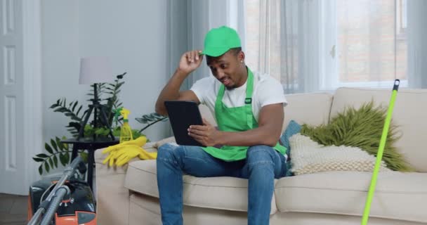 Гарний позитивний молодий впевнений чорношкірий працівник клінінгової компанії, що сидить на дивані і використовує планшетний ПК для перевірки своїх завдань при прибиранні клієнтів квартири — стокове відео
