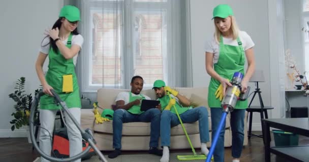 Schöne fröhliche junge gemischte Rasse Männer-Arbeiter des Reinigungsdienstes sitzen zusammen auf der Couch und bedienen den Tablet-PC, während ihre Kolleginnen im Haus der Kunden Teppiche saugen — Stockvideo