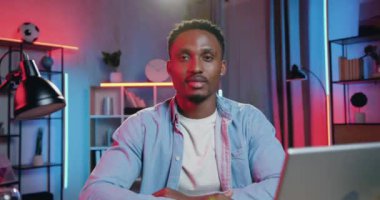 Yakışıklı, gülümseyen, mutlu, 30 yaşında, siyah tenli, iş yerinde oturan, renkli ışık efektleriyle kameraya işaret eden bir adam.