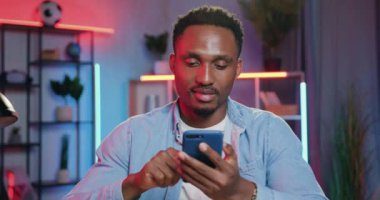 İyi görünümlü, kaygısız, genç, siyah tenli bir adamın portresi. Kameranın önünde, gece karanlığında arka planda oturuyor ve cep telefonunu kullanıyor.