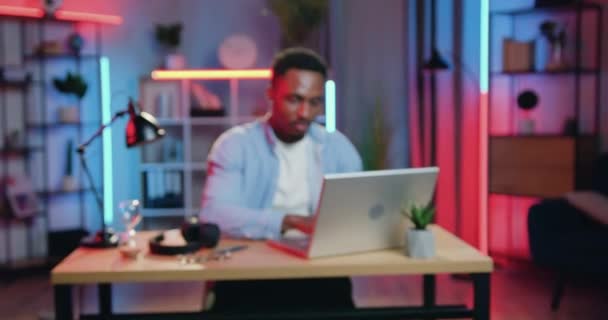 Nahaufnahme des gut aussehenden lächelnden erfolgreichen jungen schwarzhäutigen Typen, der abends zu Hause am Laptop arbeitet und mit zufriedenem Gesicht in die Kamera schaut — Stockvideo