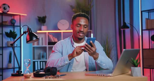 Guapo sonriente despreocupado de 30 años de edad de piel negra chico haciendo un descanso en su trabajo en el ordenador portátil en casa por la noche y charlando con amigos en el teléfono inteligente — Vídeos de Stock