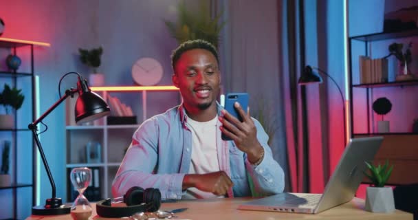 Ελκυστικός με καλή διάθεση χαμογελαστός νεαρός μαύρος τύπος απολαμβάνοντας ευχάριστη συνομιλία βίντεο στο κινητό στο σπίτι το βράδυ κατά τη διάρκεια της εργασίας του σε φορητό υπολογιστή — Αρχείο Βίντεο