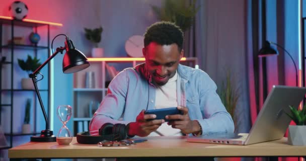 Όμορφος ενθουσιασμένος χαμογελαστός χαρούμενος νεαρός Αφρικανός Αμερικανός γιορτάζει τη νίκη στα παιχνίδια του τηλεφώνου με υψωμένα τα χέρια στο νυχτερινό φωτισμό δωματίου φόντο — Αρχείο Βίντεο