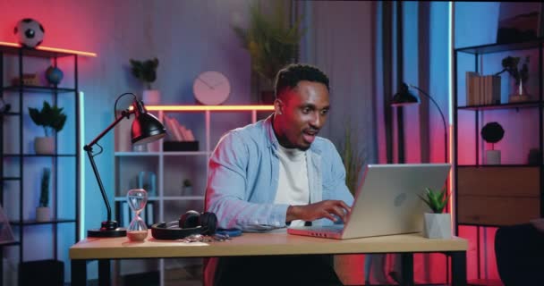 Όμορφος χαρούμενος ενθουσιασμένος νεαρός Αφροαμερικάνος κάθεται στο γραφείο μπροστά από το laptop στο δωμάτιο φωτισμού νύχτα και χαίρεται με αστεία αντίδραση όταν πάρει καλά νέα στην οθόνη — Αρχείο Βίντεο