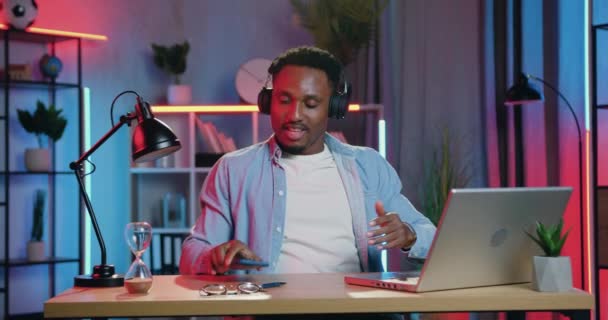 Knappe grappige vrolijke jonge zwarte huid jongen genieten van moderne muziek in hoofdtelefoon van telefoon afspeellijst tussen zijn werk op laptop thuis in de avond — Stockvideo