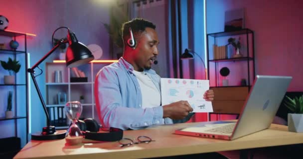 Εμφανίσιμος ικανός με αυτοπεποίθηση επιτυχημένος μαύρος τύπος σε ασύρματα ακουστικά που κάθεται μπροστά από τον υπολογιστή κατά τη διάρκεια συνάντησης βίντεο με συναδέλφους και συζητώντας κοινή επιχειρηματική στρατηγική χρησιμοποιώντας διαγράμματα — Αρχείο Βίντεο