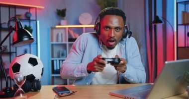 İnsan duyguları konsepti. Yakışıklı, mutlu, kulaklıklı, siyah tenli genç adam akşamları evdeki video oyunlarında kazandığı zaferin tadını çıkarıyor.