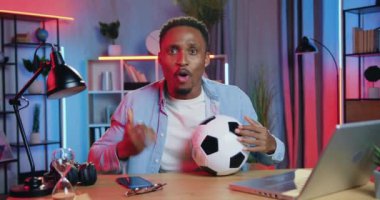 Yakışıklı, gülümseyen, heyecanlı, siyah tenli bir erkek futbol fanatiğinin portresi. Futbol maçını izlerken kameranın önünde oturan ve golü atan