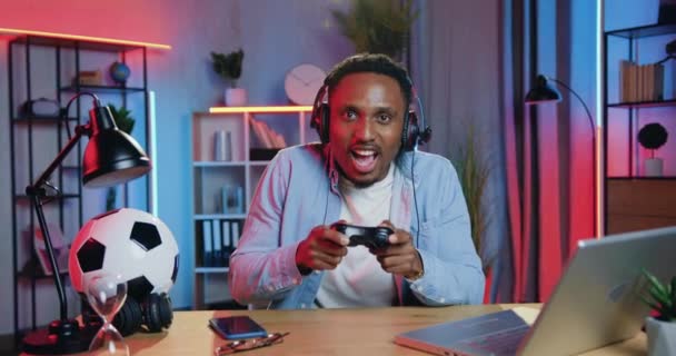 Human reactions concept where good-looking excited funny young black-skinned guy in headphones sitting in front of camera and playing video games using gamepad and is dissatisfied as he lost the game — Stock Video