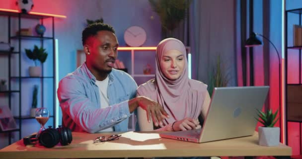 Bonito cara positivo de pele preta discutindo projeto conjunto, juntamente com mulher árabe bonita em hijab sentado na frente do computador no escritório da noite — Vídeo de Stock