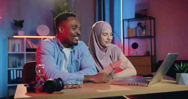 Good-loooking souriant satisfait homme afro-américain et femme musulmane dans le hijab assis devant l'ordinateur du bureau de nuit pendant le chat vidéo avec freinds — Video