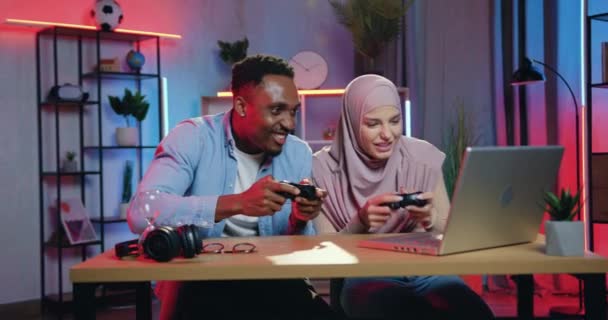 Adorable heureux souriant afro-américain gars et fille musulmane dans hijab jouer à des jeux vidéo sur ordinateur à l'aide de manettes et célébrer la victoire avec cinq dans la soirée — Video