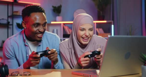 Atraente feliz engraçado sorrindo cara de pele preta comemorando a vitória no jogo de vídeo sobre seu parceiro fêmea árabe durante competições no computador no escritório em casa à noite — Vídeo de Stock