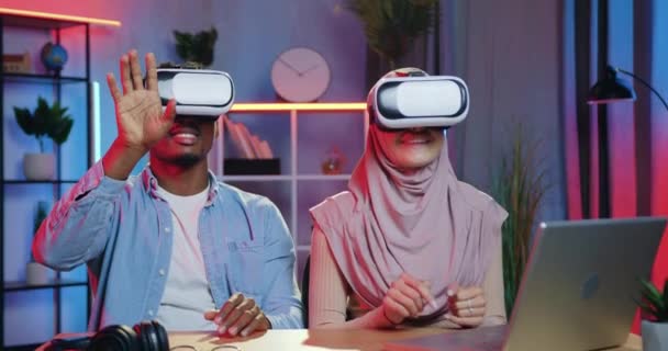 Pareja de raza mixta inteligente positiva segura agradable sentada delante de la cámara en gafas 3D especiales y manos móviles en la pantalla virtual trabajando por la noche en la oficina, cámara lenta — Vídeo de stock