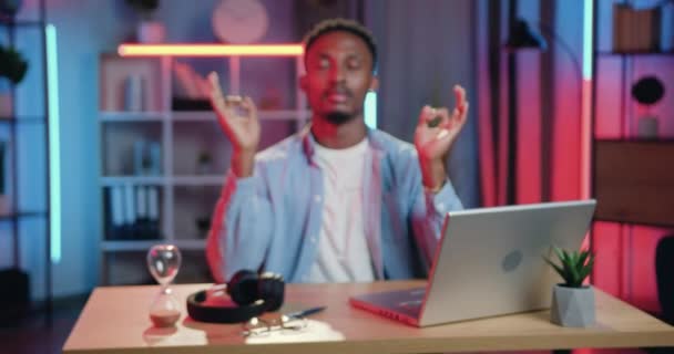 Handsome peaceful young african american sitting at his workplace at home in the evening and meditating with closed eyes and folded in mudra gesture hands — Stock Video